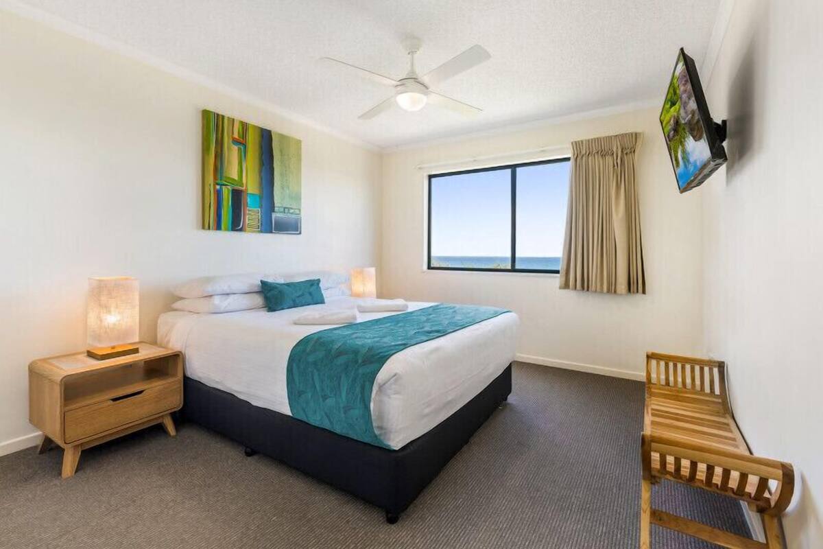 A hotel room with a bed, a nightstand, a desk, a chair, a ceiling fan, a window with curtains, and a painting on the wall.