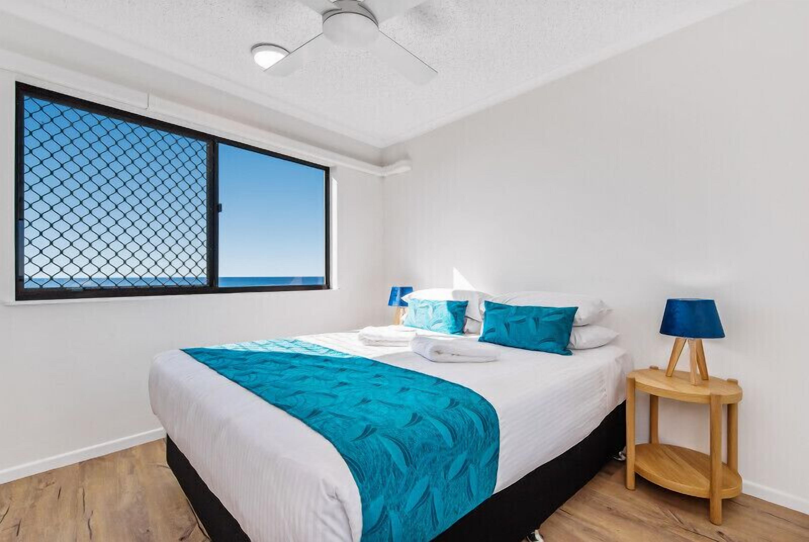 A bedroom with a bed, a window with a view, a ceiling fan, and a bedside table with a lamp.