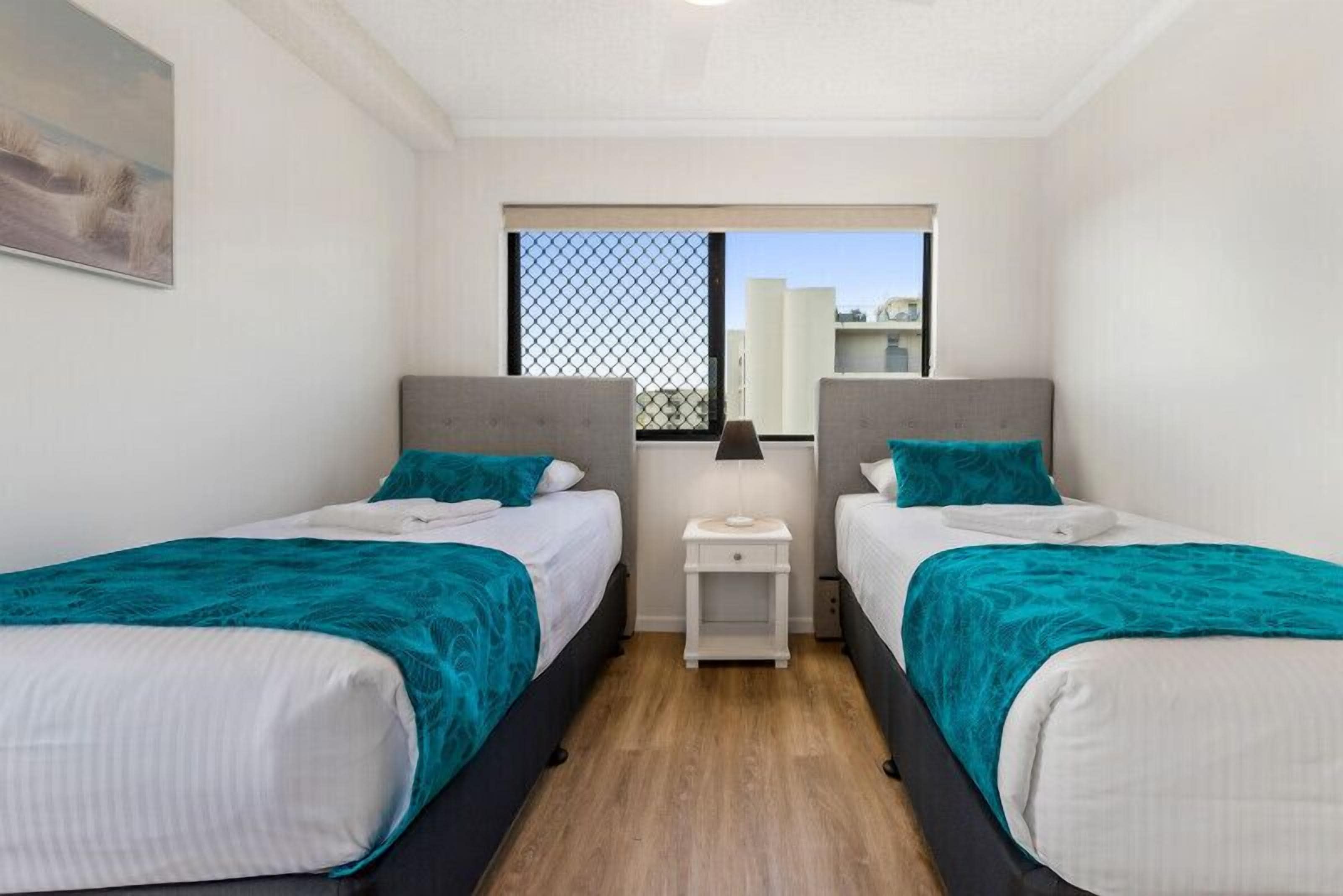A bedroom with two beds, each with a teal bedspread, a small bedside table with a lamp, and a window with a view of buildings.