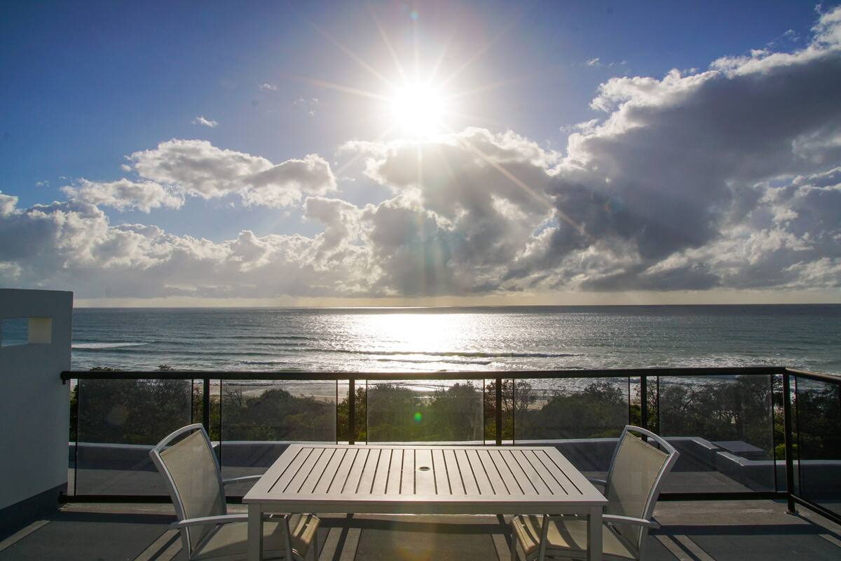 A balcony with a table and chairs, overlooking the ocean with the sun shining through the clouds.