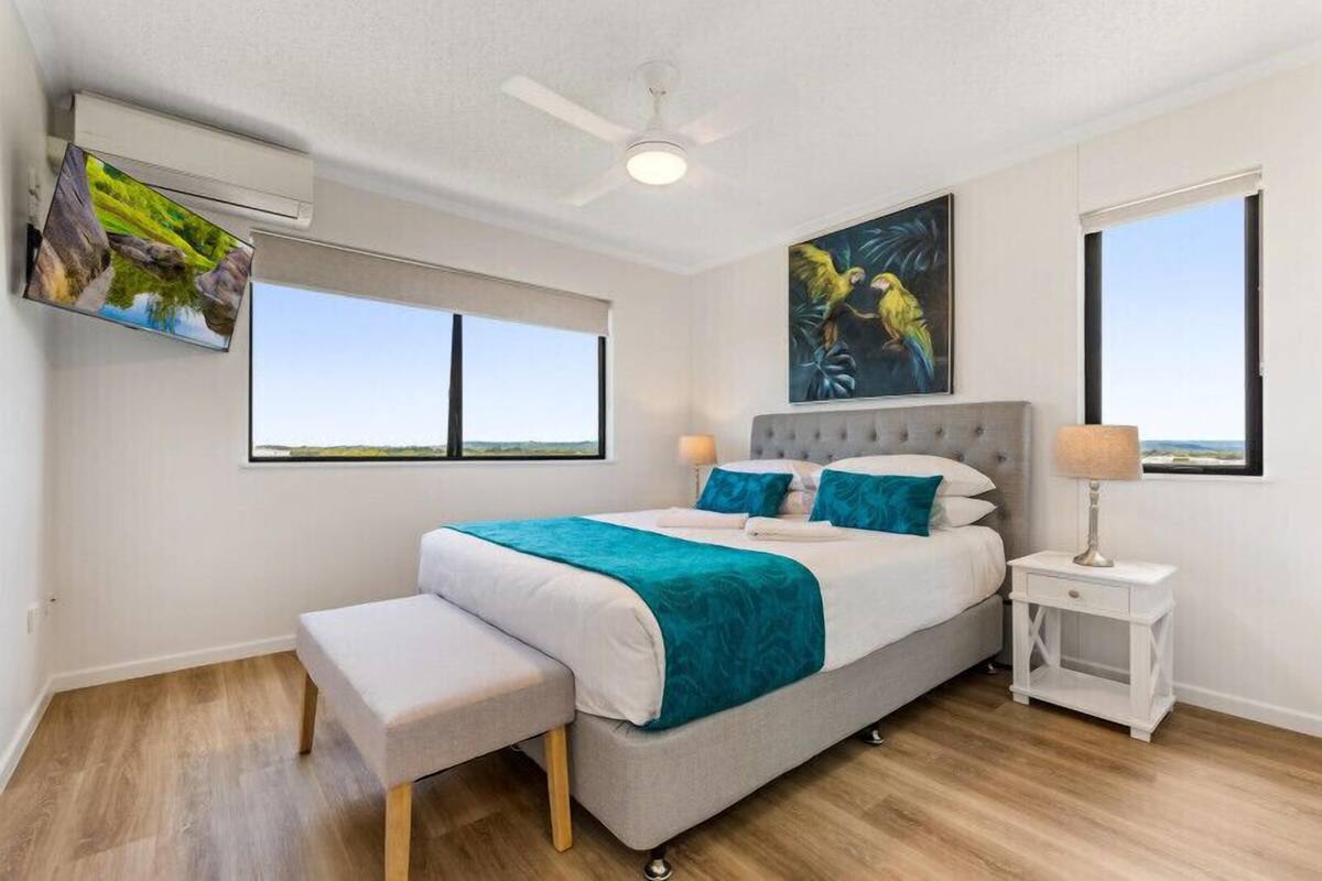 A modern bedroom with a bed, a bench, a nightstand, a ceiling fan, and a TV mounted on the wall.