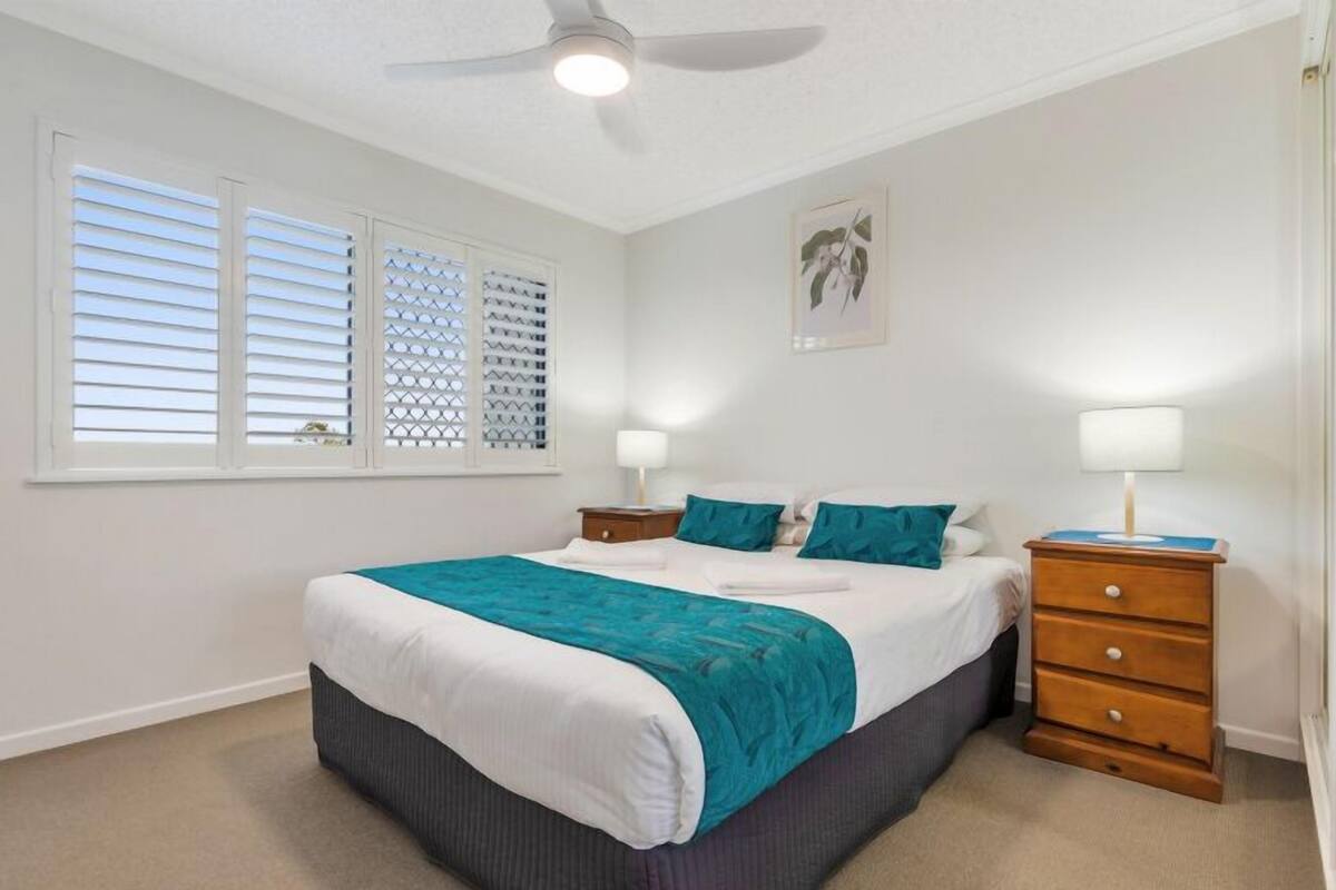 A bedroom with a bed, two nightstands, a ceiling fan, and a window with blinds.