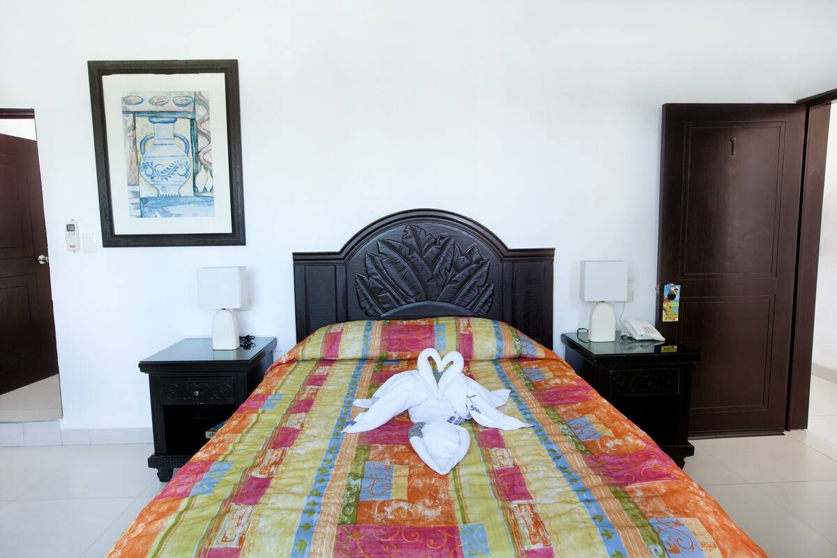 A neatly made bed with a colorful quilt and a white towel shaped like a swan on the center. There are two bedside tables with lamps and a framed artwork on the wall.