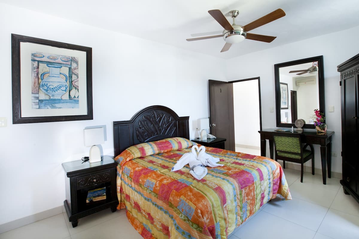 A bedroom with a bed, a desk, a chair, a nightstand, a ceiling fan, a framed picture, and a mirror.