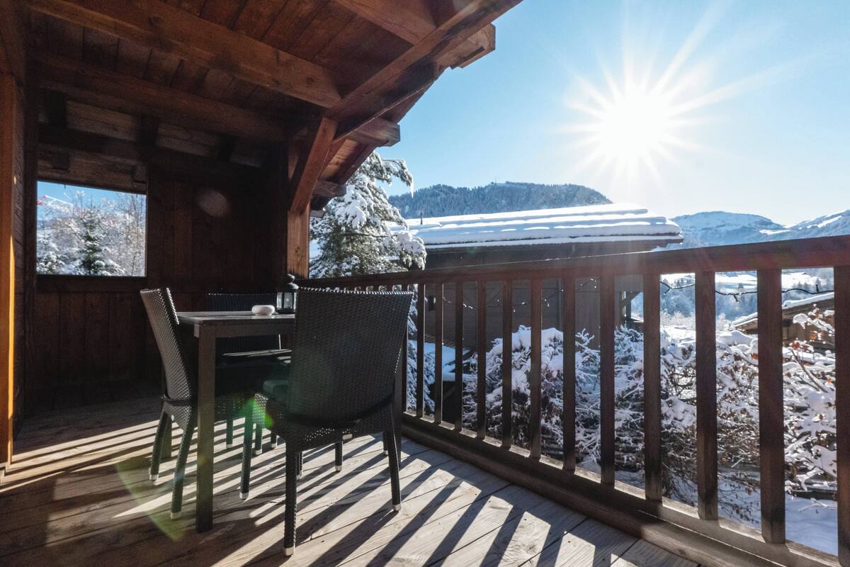 A wooden deck with a table and chairs, overlooking a snowy landscape with mountains in the background.