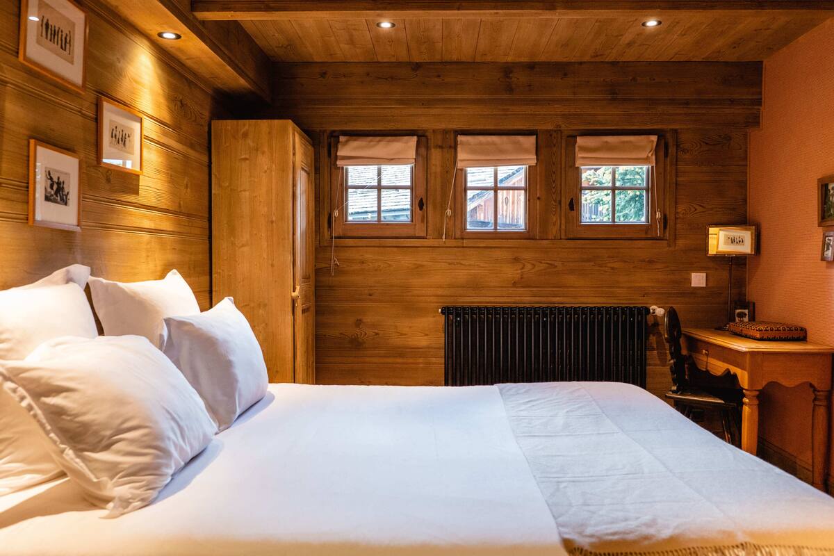 A wooden-paneled bedroom with a bed, two windows with curtains, a radiator, and a small table with a chair.