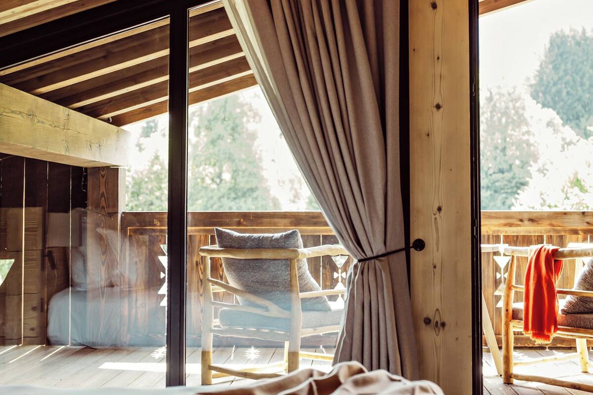 A wooden cabin interior with a bed, large windows, and a view of the outdoors.