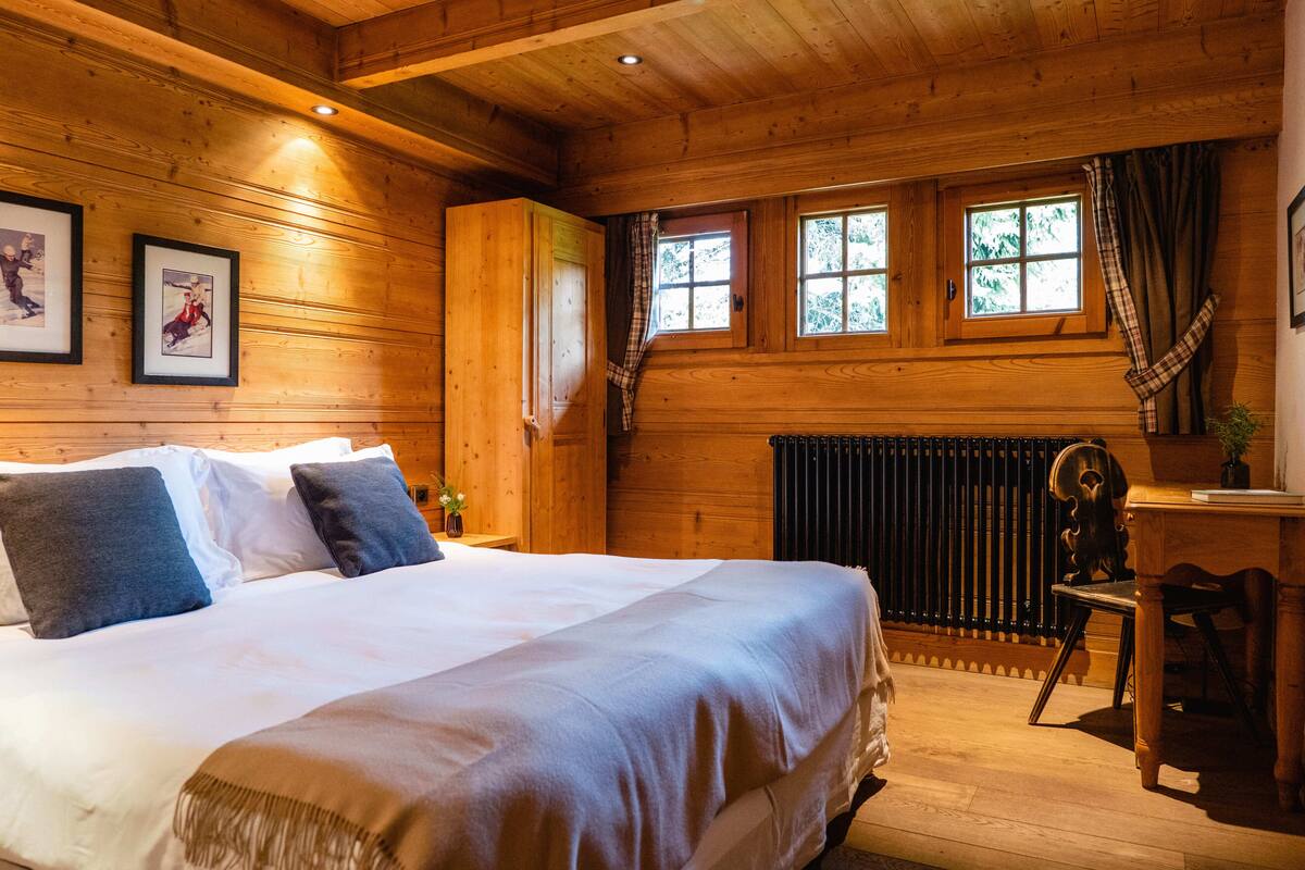 A wooden cabin room with a bed, two windows, a radiator, and a chair.