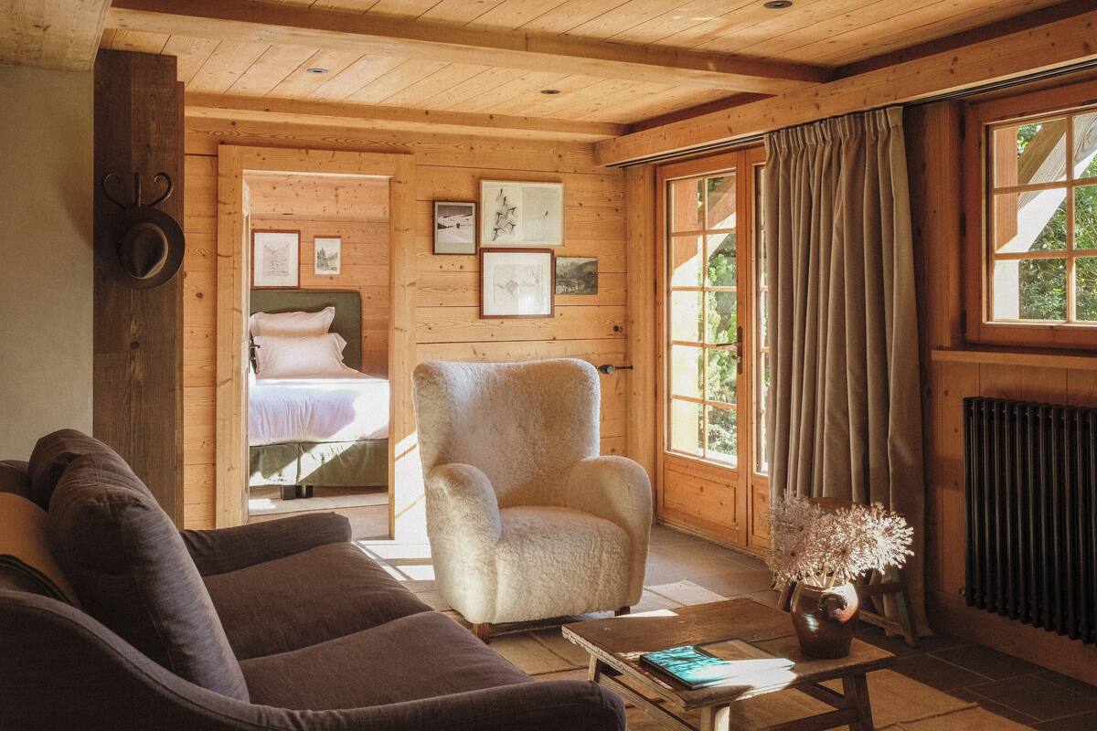 A cozy room with a wooden ceiling, a large window with curtains, a bed, a plush armchair, and a coffee table with a vase.