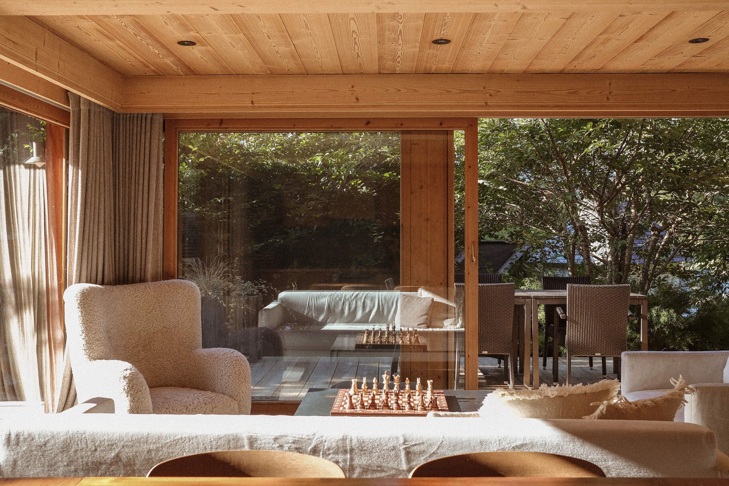 A cozy room with a large window, a comfortable chair, a small table with a chessboard, and wicker furniture.