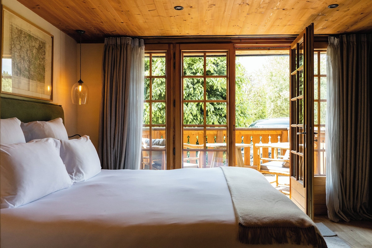 A large bed with white linens, a wooden headboard, and a view of an outdoor deck with wooden furniture.