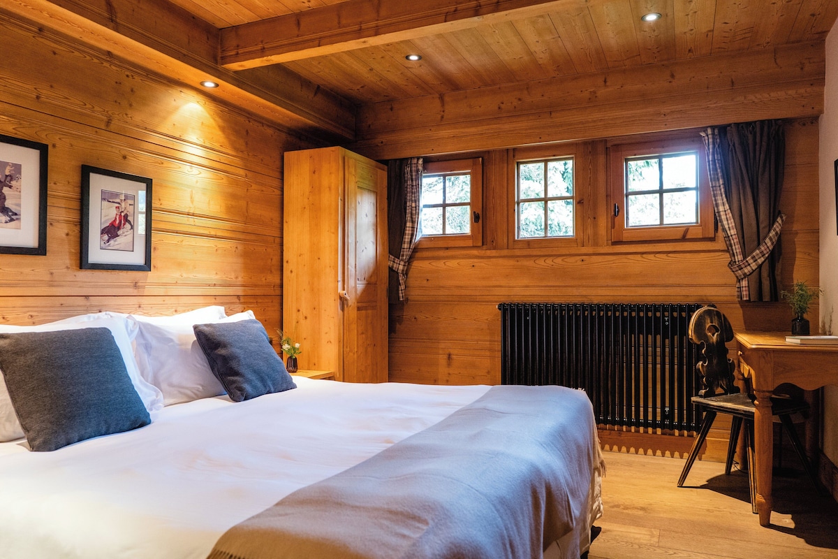 A wooden cabin room with a bed, two framed pictures on the wall, a desk with a chair, and a window with curtains.