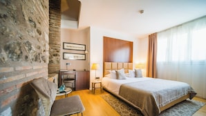 Una habitación de hotel con pared de ladrillo, una cama con cabecera de madera, un escritorio con una silla y una mesita con una lámpara.