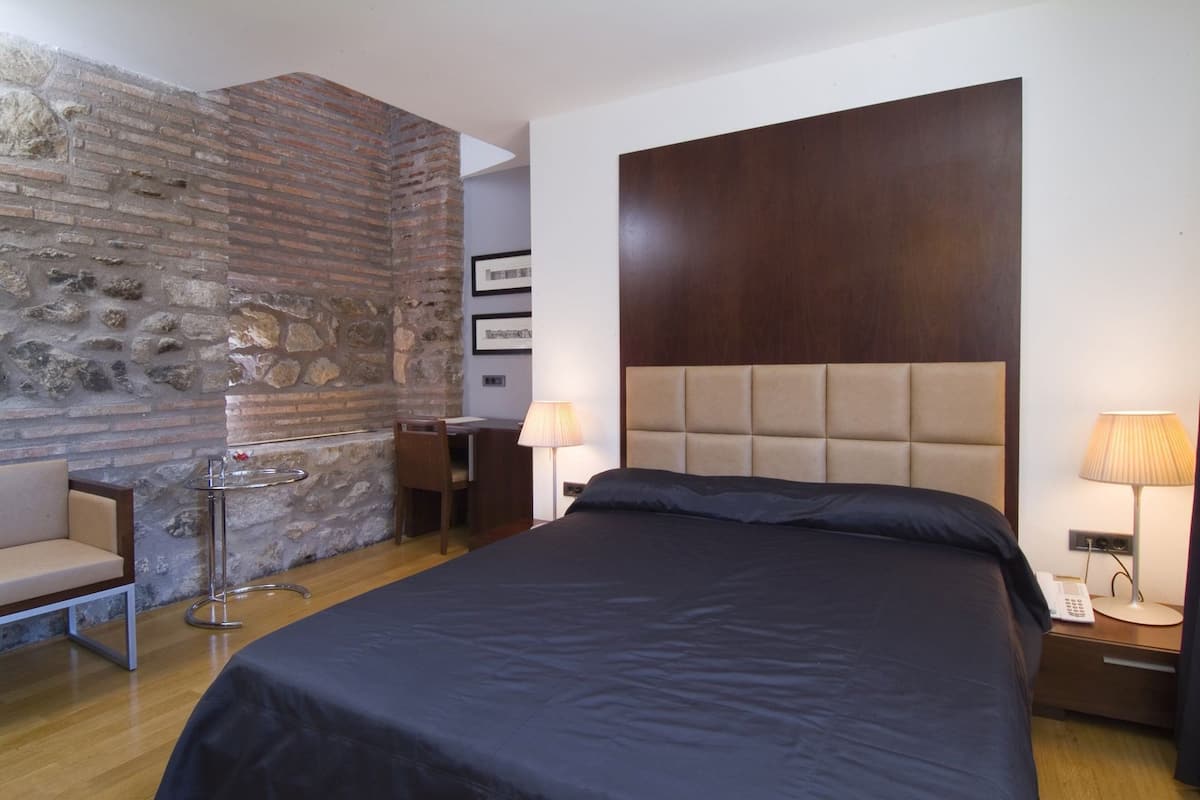 A hotel room with a bed, a desk, a chair, a lamp, and a stone wall.