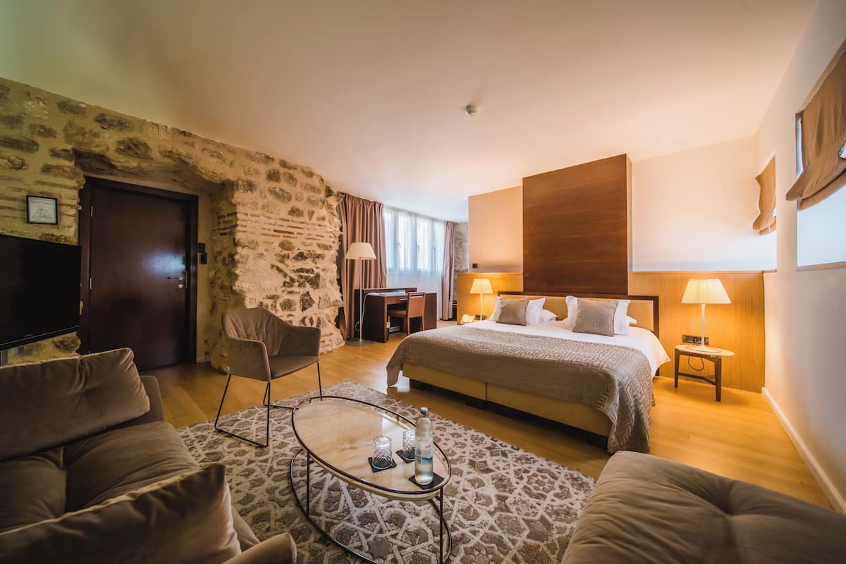 A hotel room with a stone wall, a bed with a wooden headboard, a glass coffee table, a sofa, and a desk with a chair.