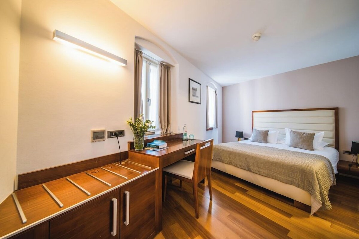 A bedroom with a bed, bedside table, desk, chair, and a window with curtains.
