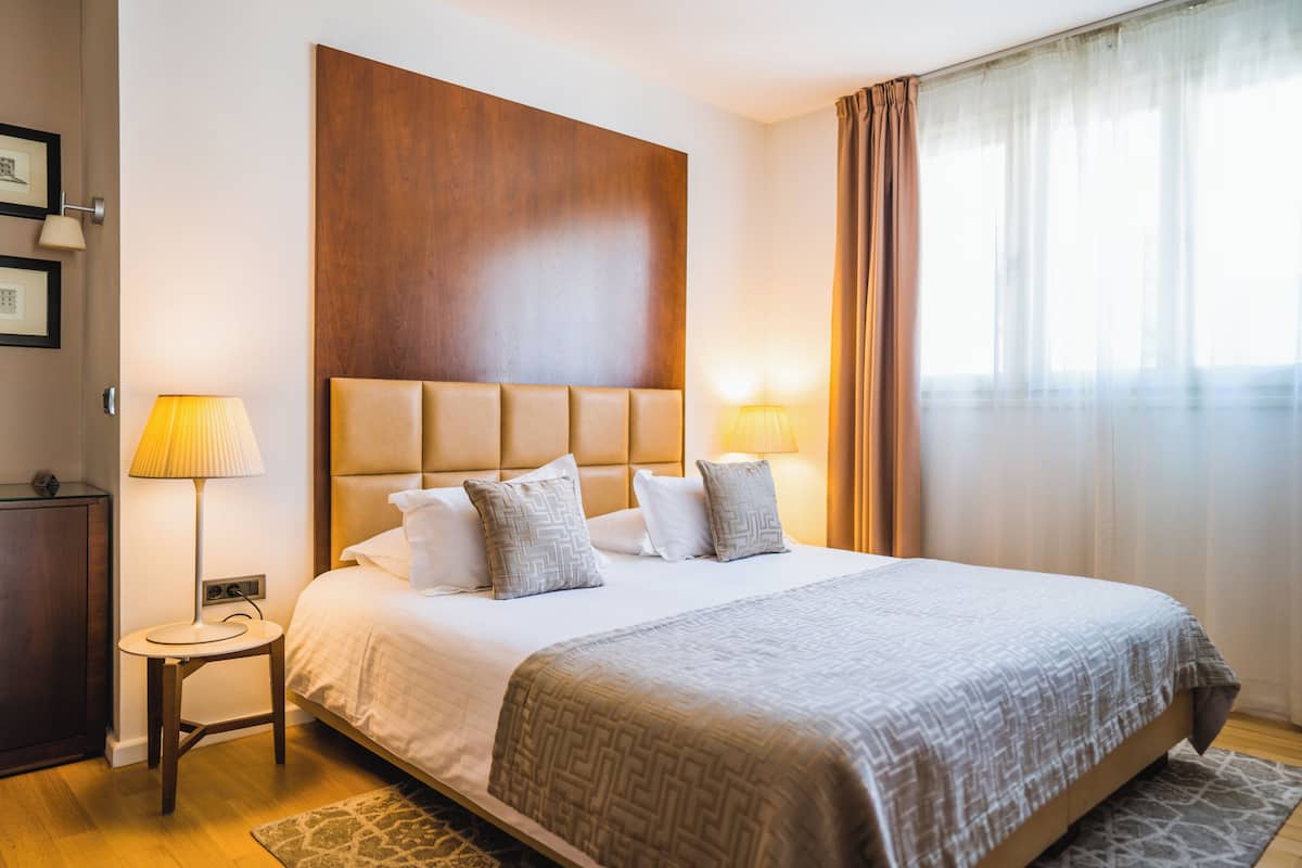 A hotel room with a large bed, two bedside lamps, a wooden headboard, a nightstand, and a large window with curtains.