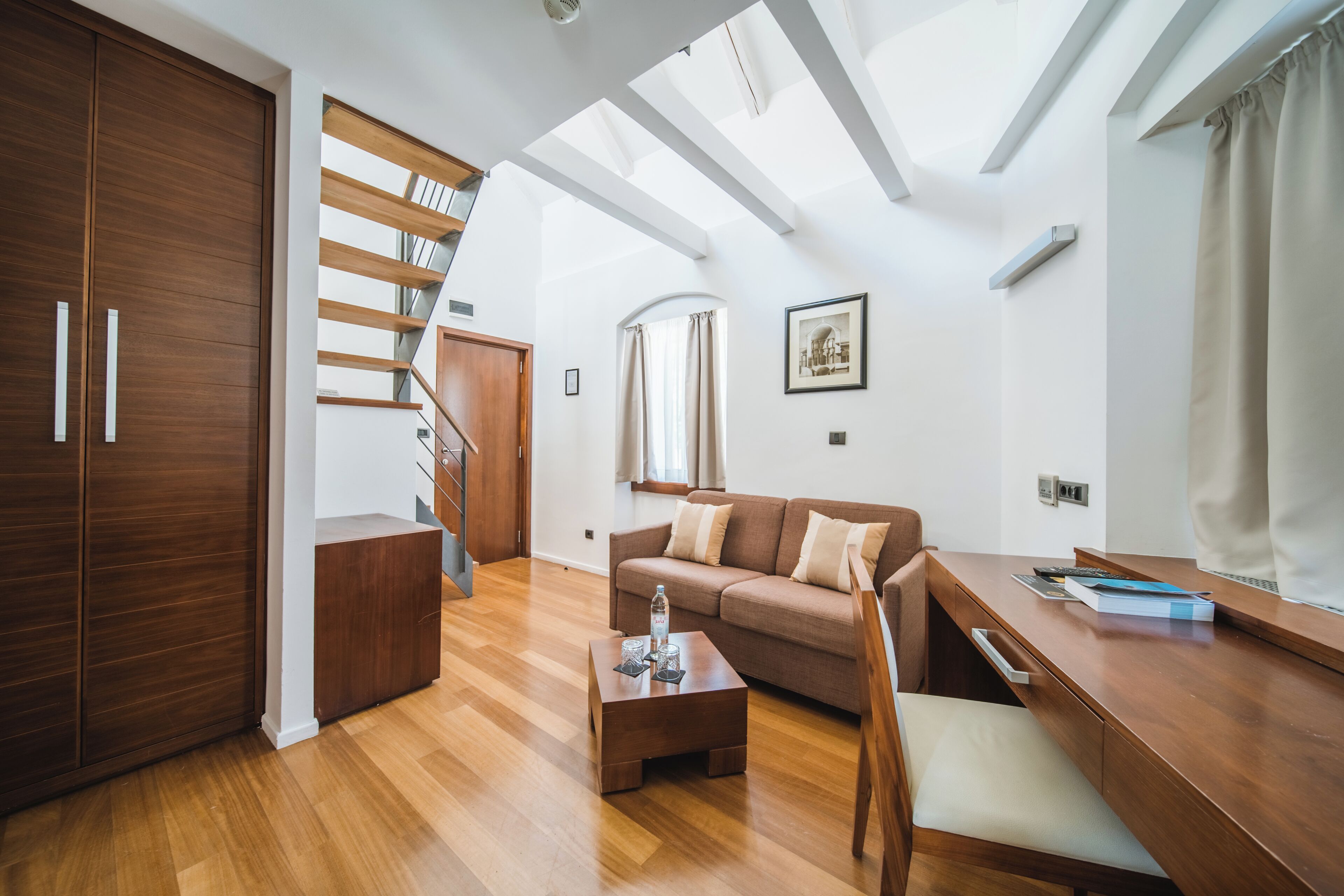 Un salon moderne avec un plancher en bois, un canapé, une table basse et un bureau. Un escalier mène à un niveau supérieur.