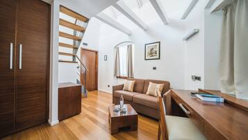 A modern living room with a wooden floor, a sofa, a coffee table, and a desk. There is a staircase leading to an upper level.