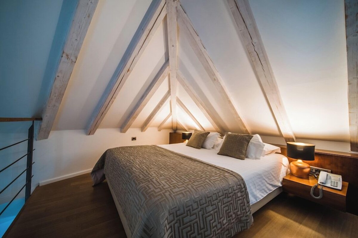 A bedroom with a sloped ceiling, a bed with a patterned bedspread, a bedside table with a lamp, and a telephone.