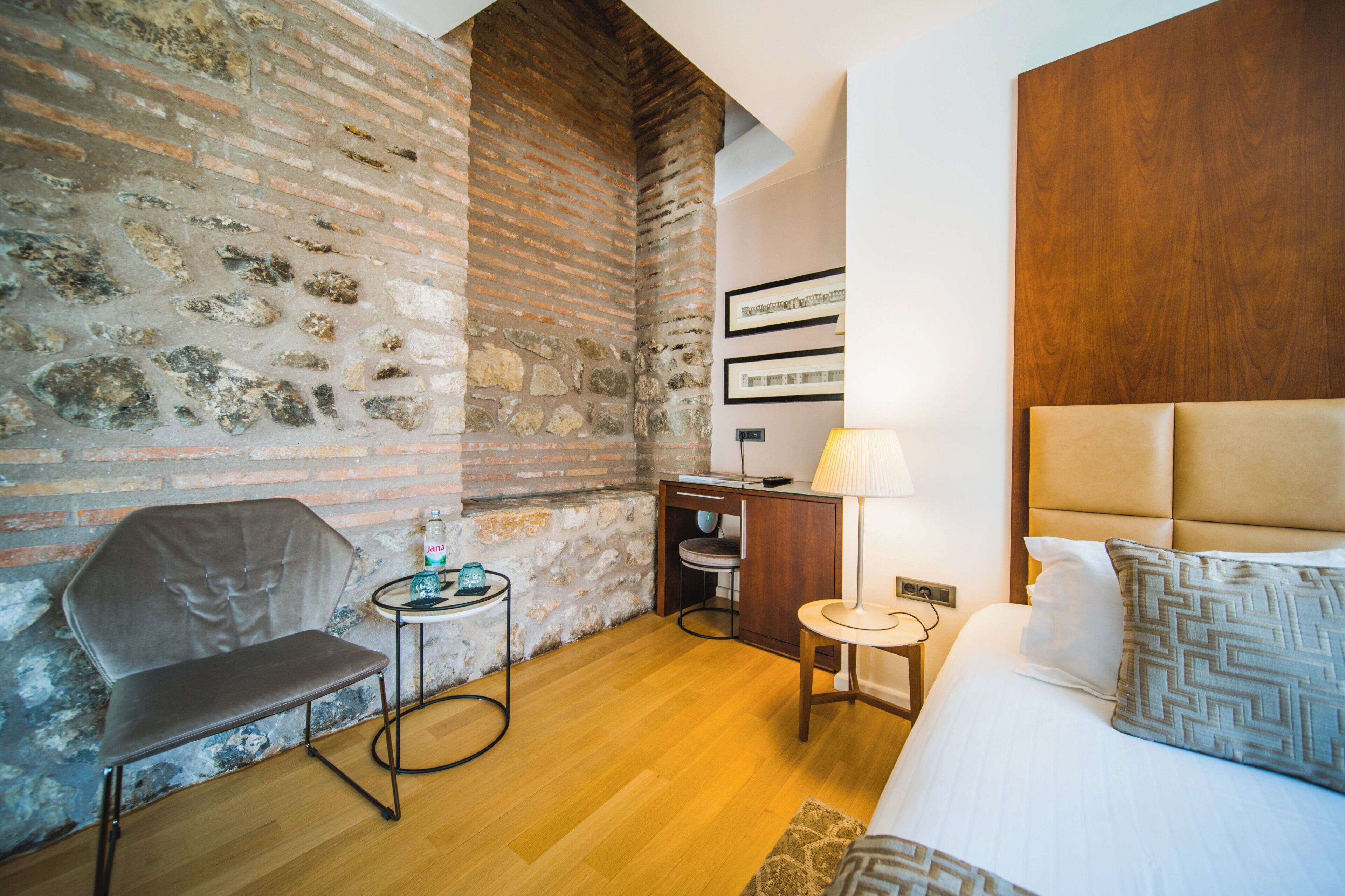 Ein Hotelzimmer mit Backsteinwand, einem Bett mit Holz Kopfteil, einem Schreibtisch mit Lampe, einem Sessel und einem kleinen Tisch ebenfalls mit Lampe.
