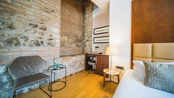 Ein Hotelzimmer mit Backsteinwand, einem Bett mit Holz Kopfteil, einem Schreibtisch mit Lampe, einem Sessel und einem kleinen Tisch ebenfalls mit Lampe.