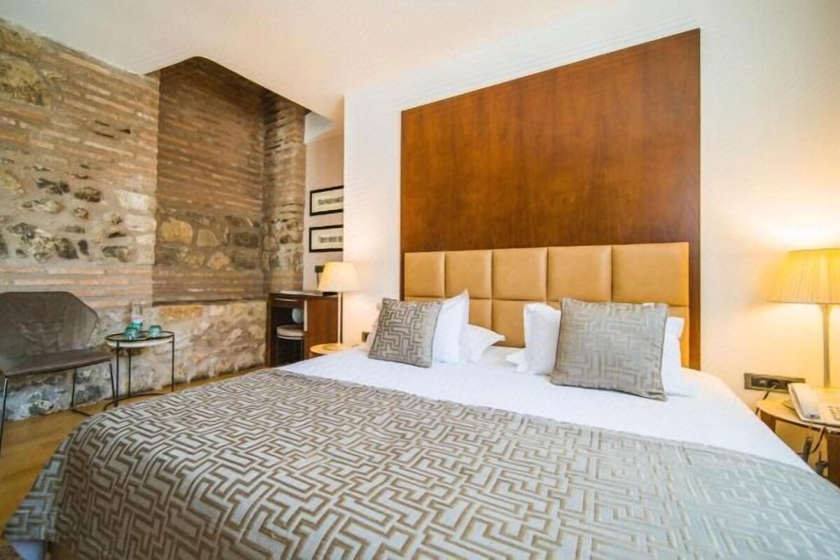 A hotel room with a large bed, a stone wall, a wooden headboard, and a small table with a lamp.