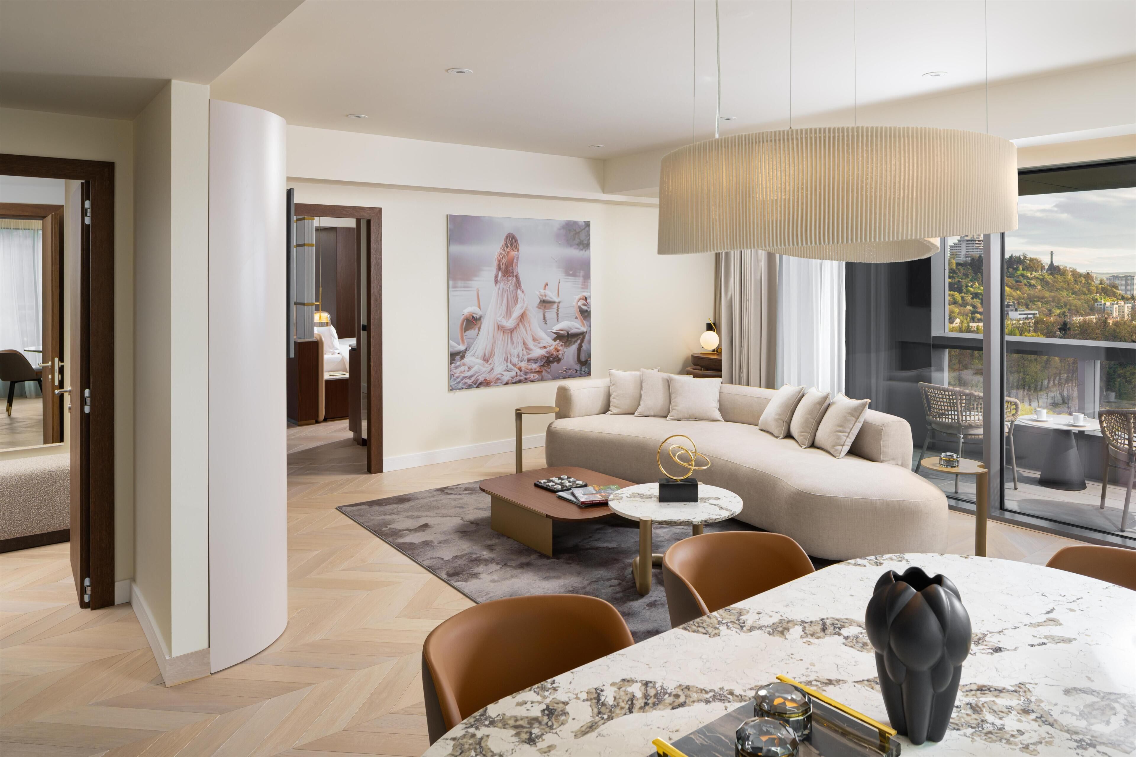 A modern hotel room with a large window, a sofa, a dining table, and a painting on the wall.