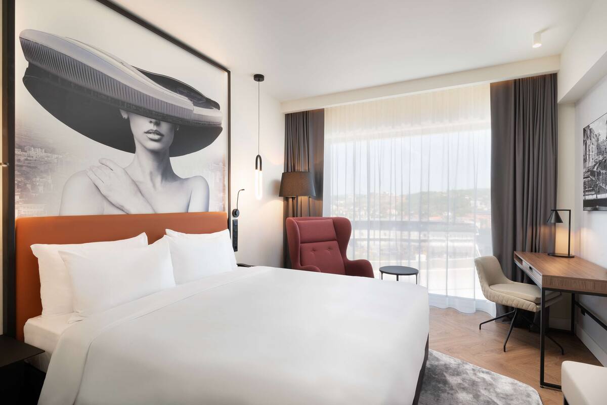 Quarto de hotel com uma cama grande, uma escrivaninha, uma cadeira e vista para a cidade pela janela.