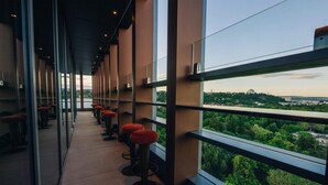 Un pasillo con ventanas altas, taburetes rojos y vistas a un paisaje urbano.
