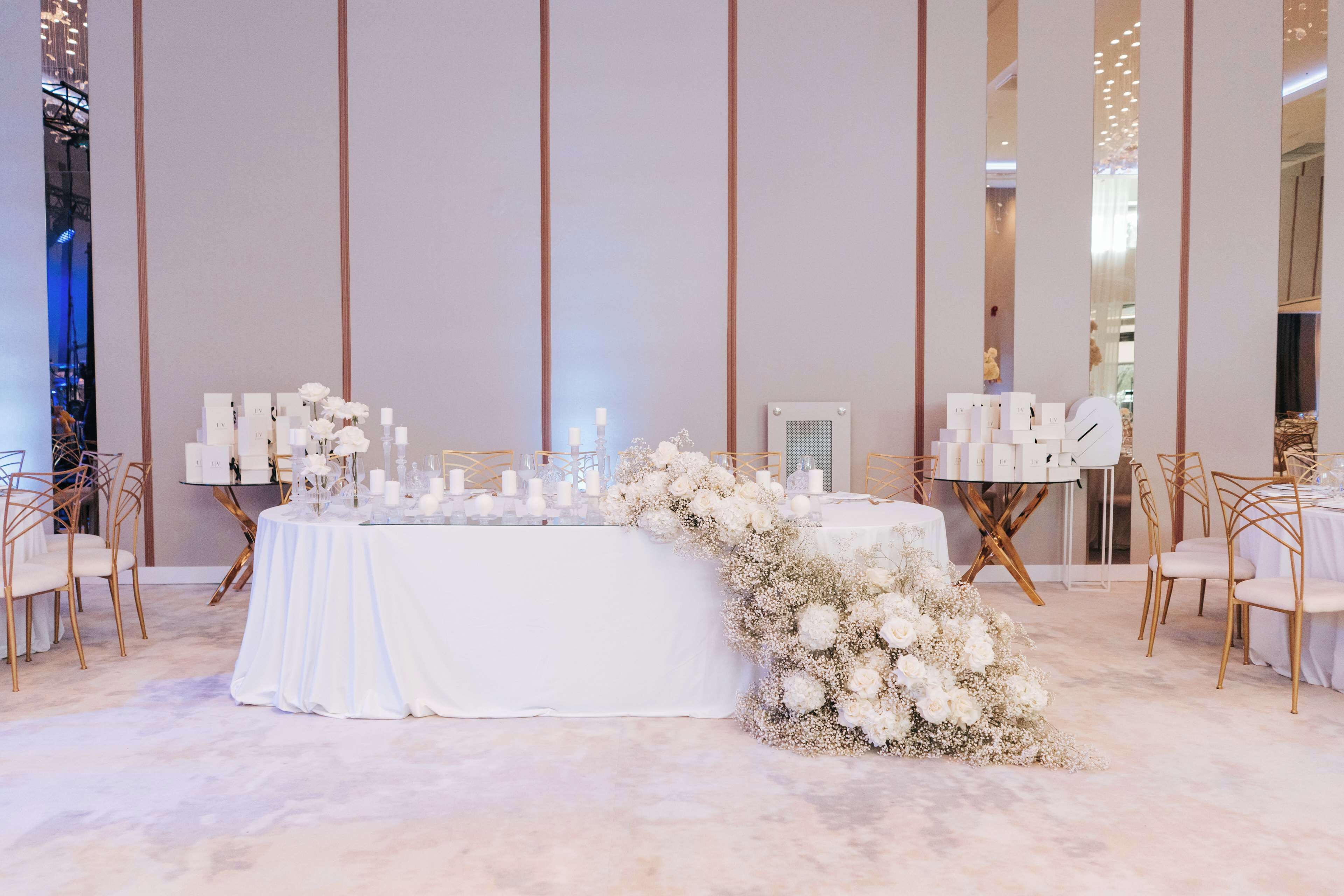 A wedding reception setup with a white table, floral arrangements, and gold chairs.