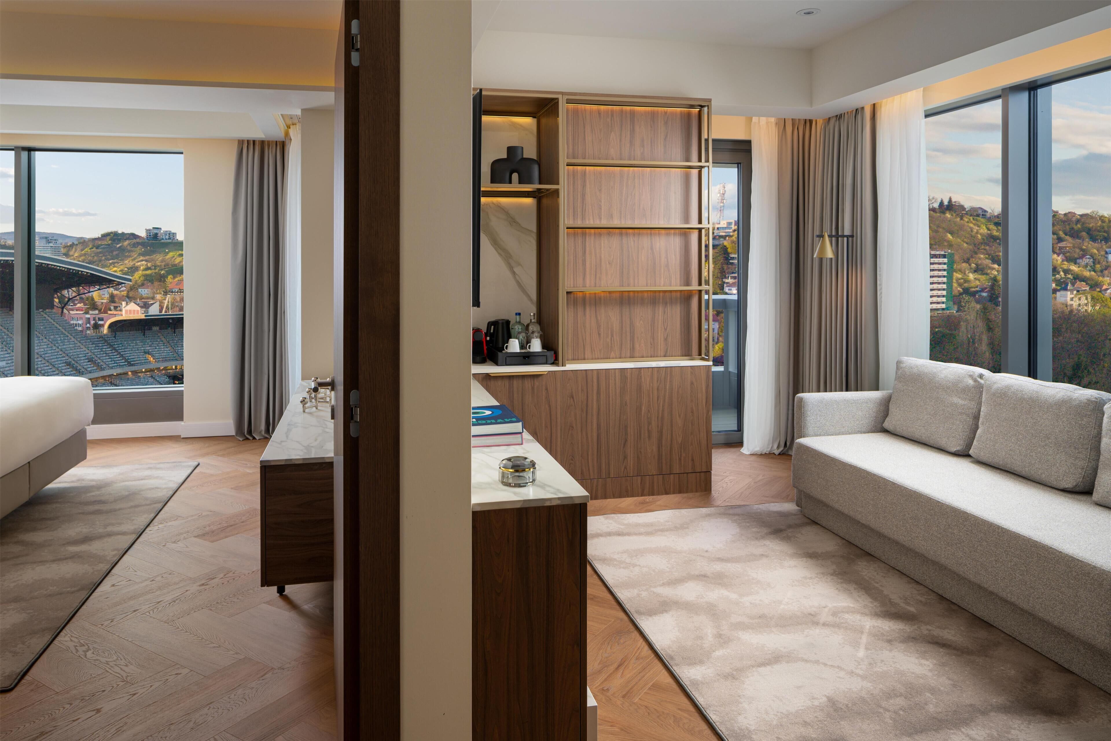 A modern hotel room with a large window, a sofa, a desk, and a built-in wardrobe.