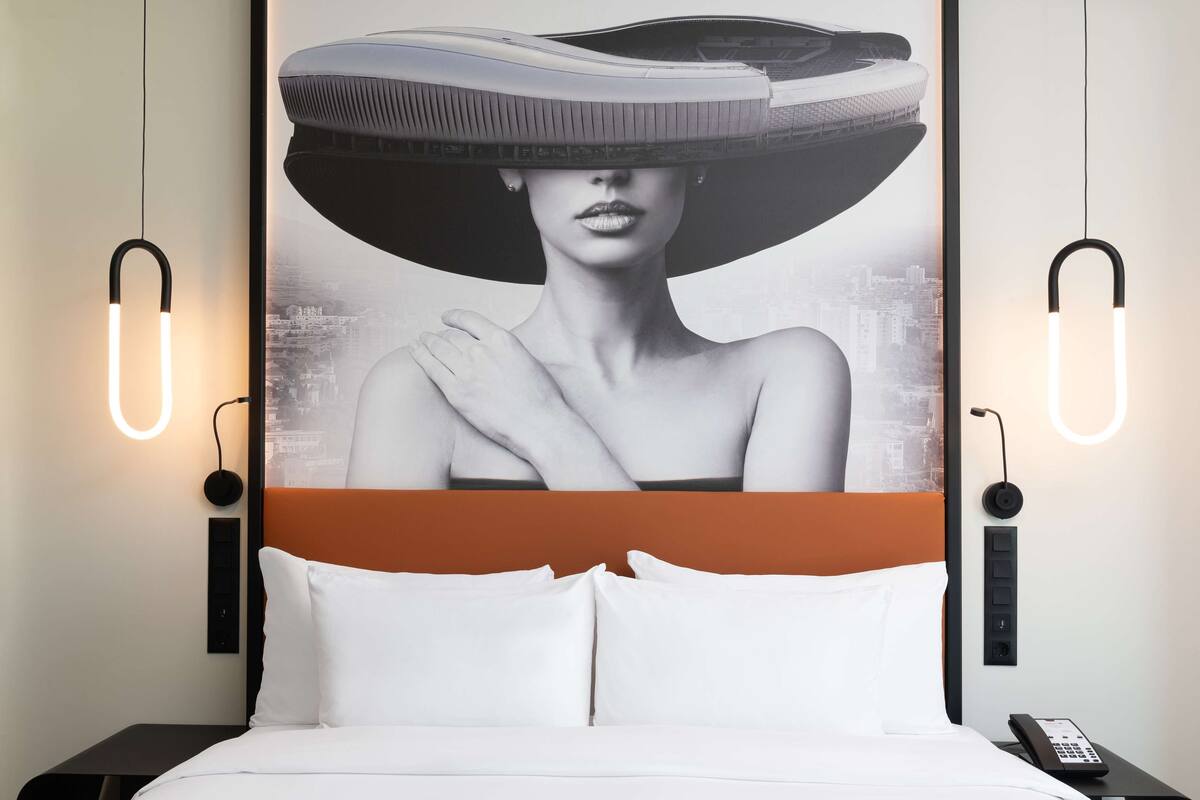 Quarto de hotel moderno com uma cama grande, um cartaz de paisagem urbana e duas luminárias fixadas na parede.