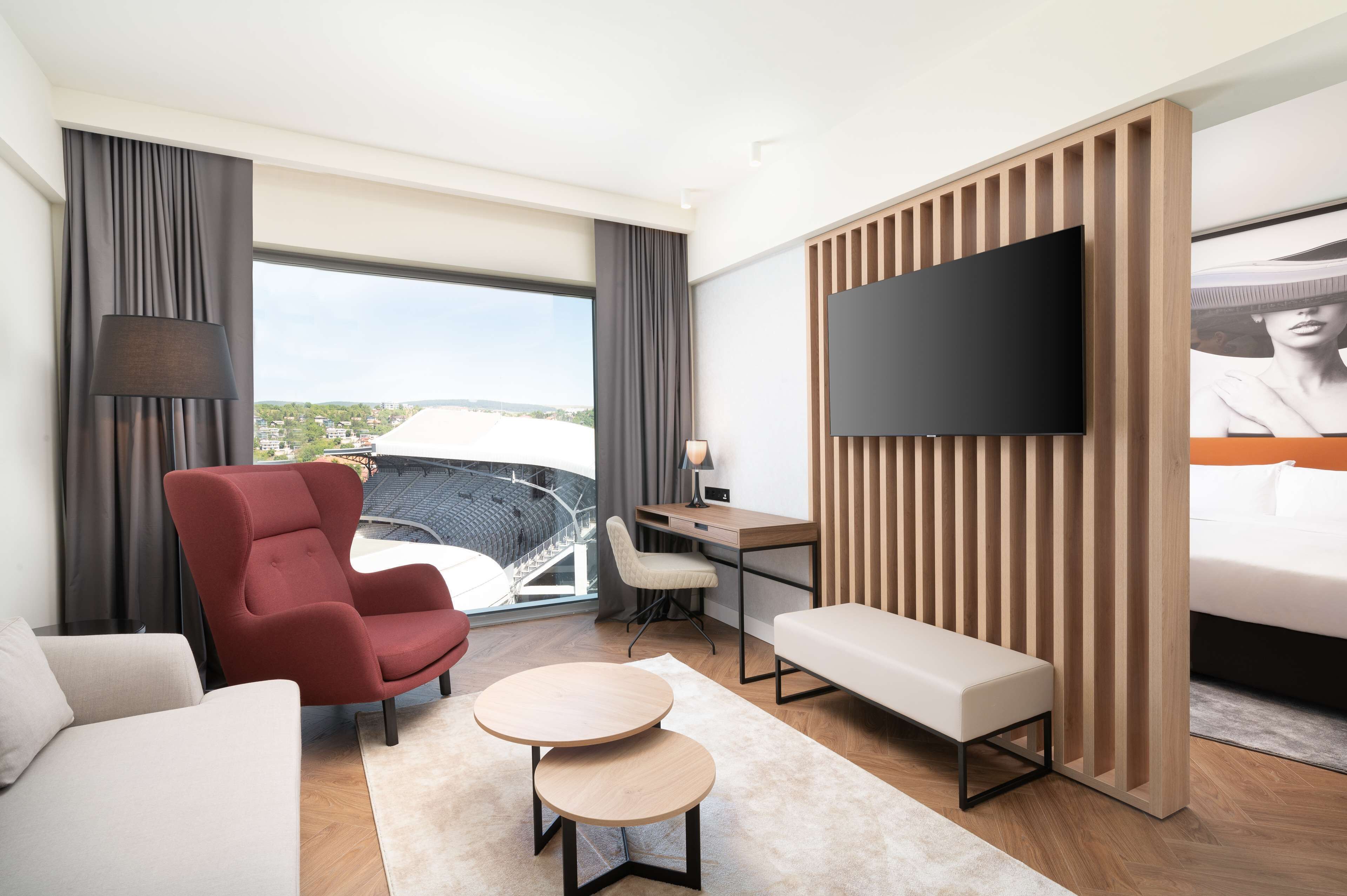 Een moderne hotelkamer met een groot raam, een rode fauteuil, een bureau, een televisie en een bed.