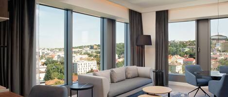 Una sala de estar moderna con un sofá, dos mesitas y vistas a un paisaje urbano a través de amplias ventanas.