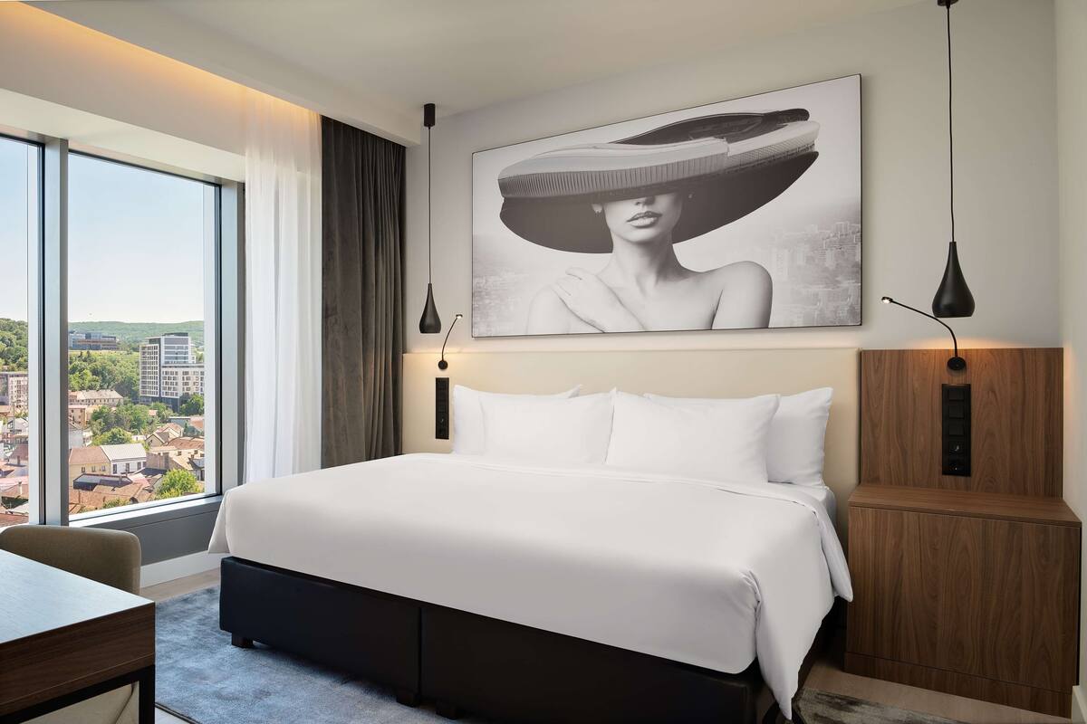 Quarto de hotel moderno com uma cama grande, uma escrivaninha e vista para a cidade através de grandes janelas.