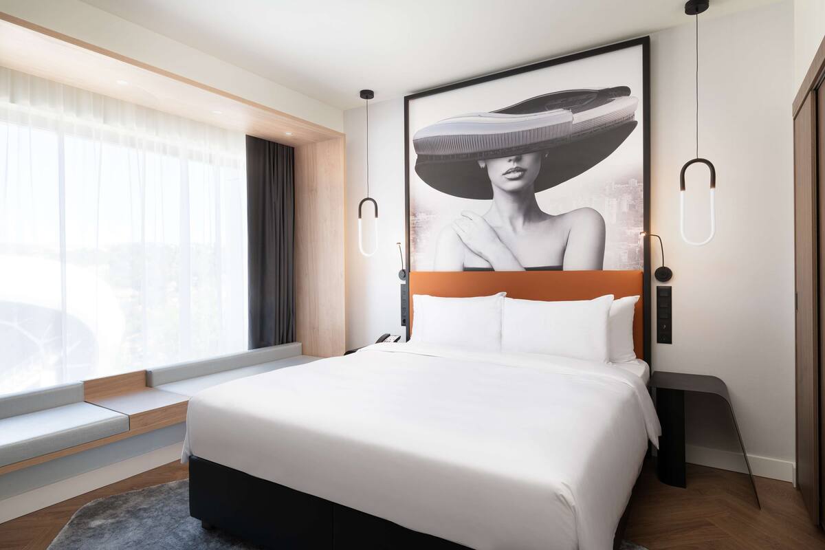 Quarto de hotel moderno com uma cama grande, um cartaz de paisagem urbana, uma janela com vista e uma mesinha de cabeceira.