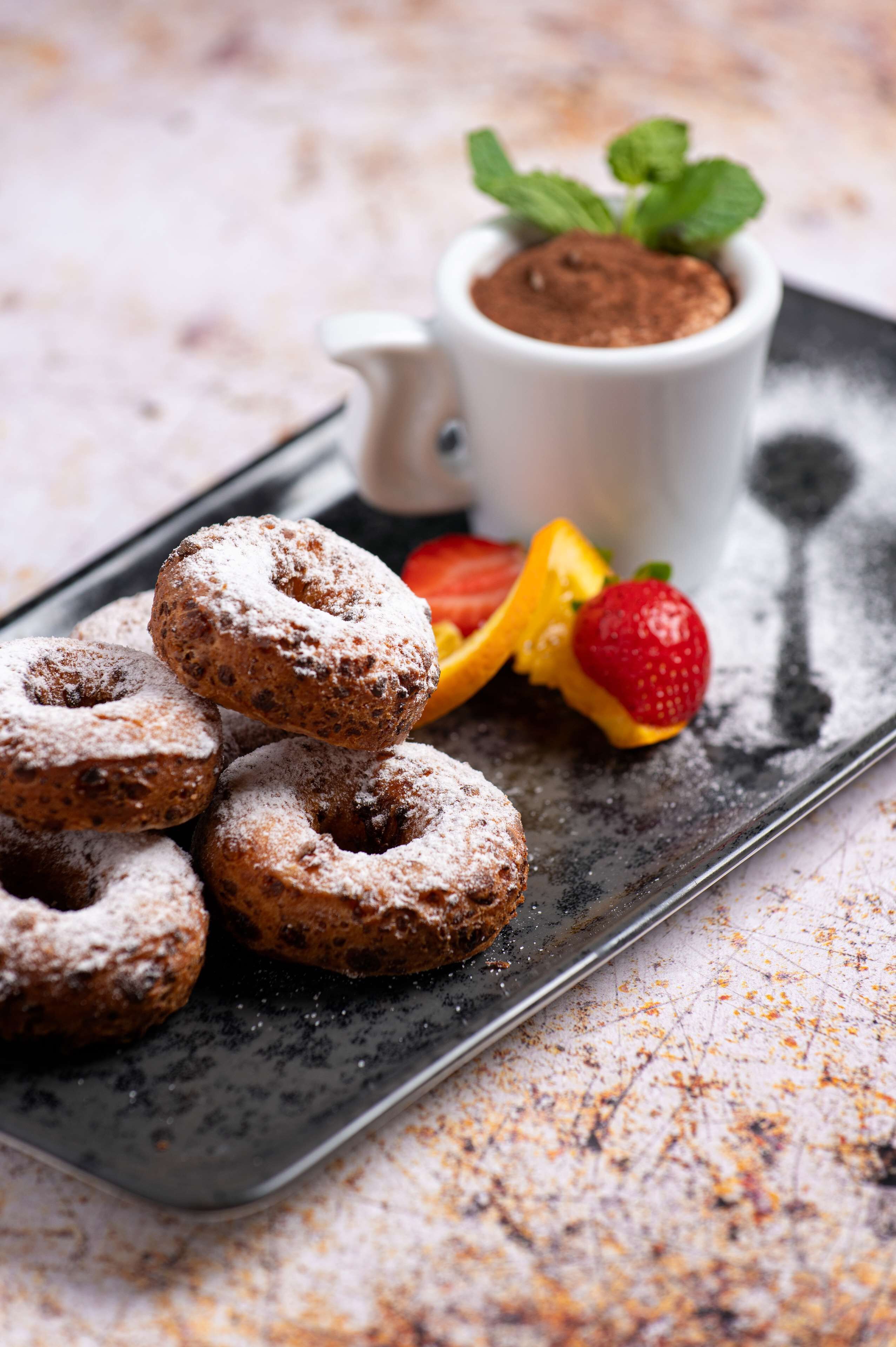 A tray of powdered donuts with a small cup of cocoa powder and some fruit.
