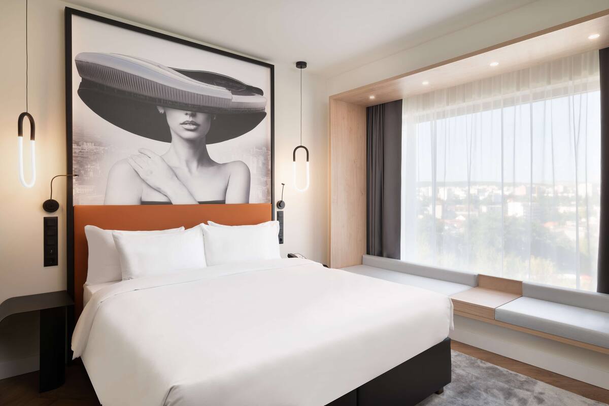 Quarto de hotel moderno com uma cama grande, vista da cidade pela janela e uma obra de arte emoldurada acima da cama.