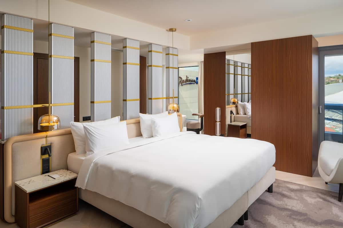 Quarto de hotel moderno com uma cama grande, mesas de cabeceira, uma escrivaninha e vista para a cidade.