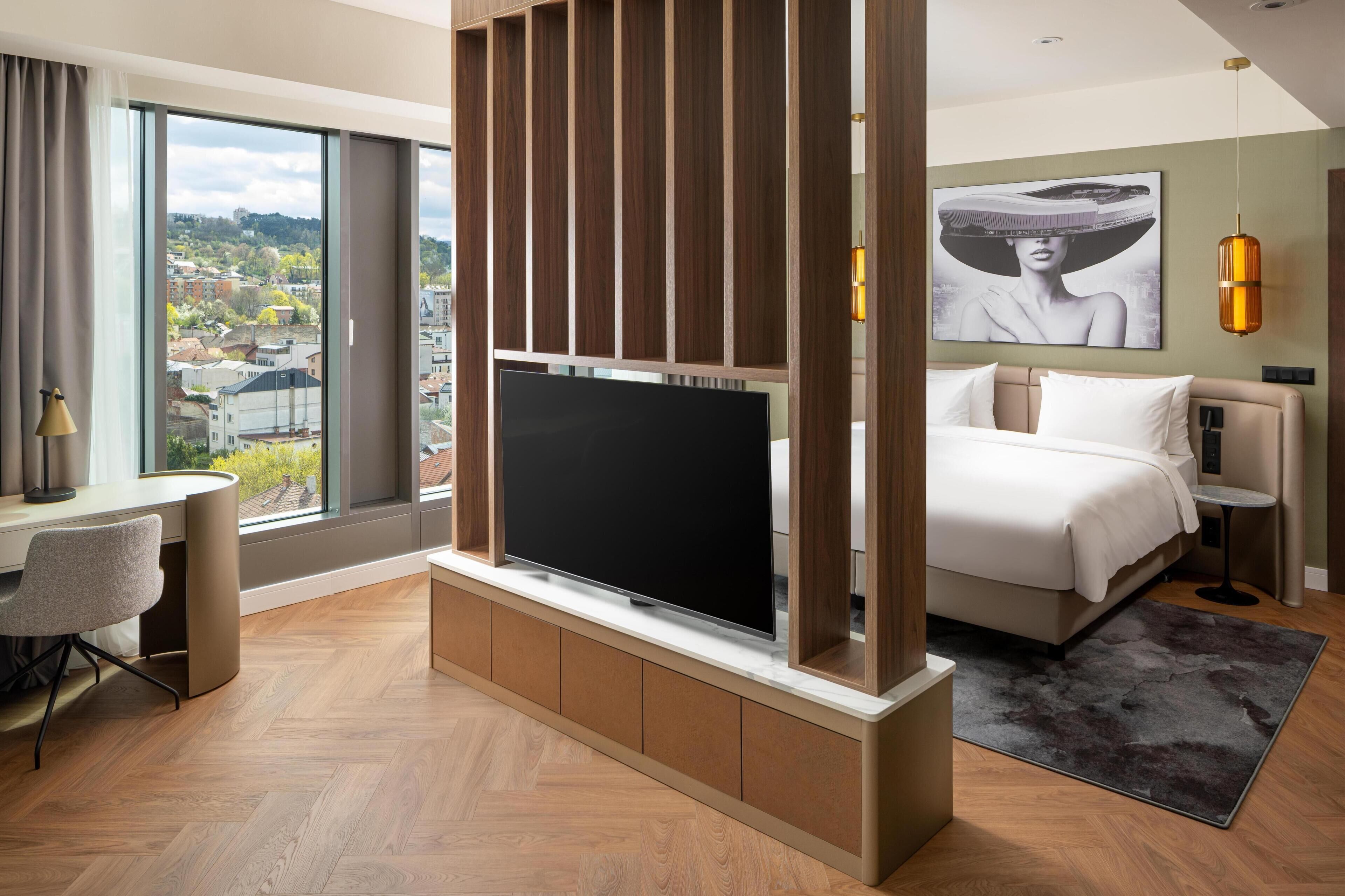 Habitación de hotel moderna con una televisión grande, una cama, un escritorio y vistas a la ciudad.