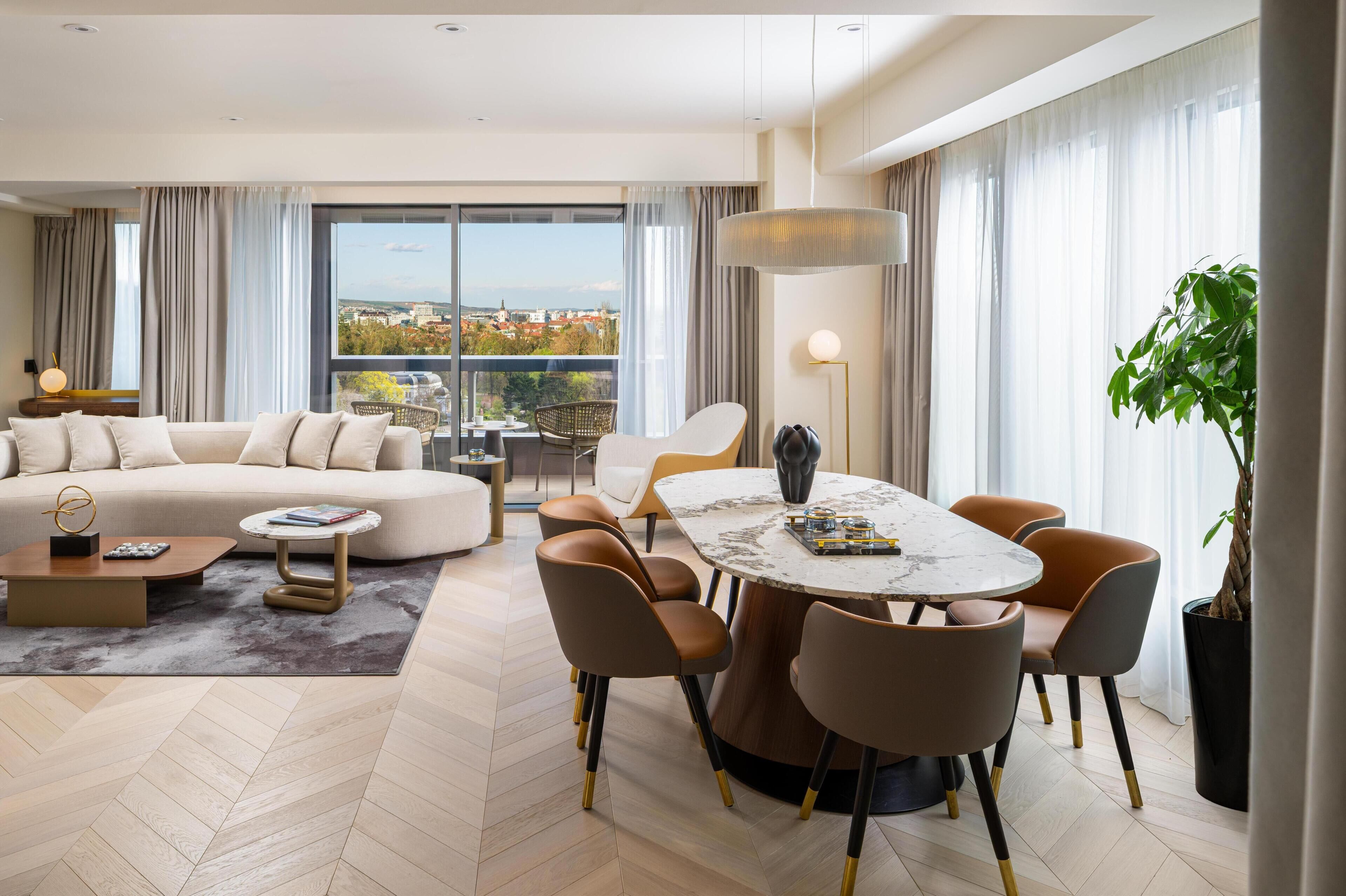 A modern living room with a large sofa, a dining table with chairs, a potted plant, and a view of the city through large windows.