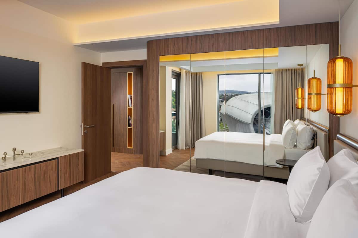 Quarto de hotel com uma cama grande, TV na parede, aparador de madeira com espelho e vista para a cidade pela janela.