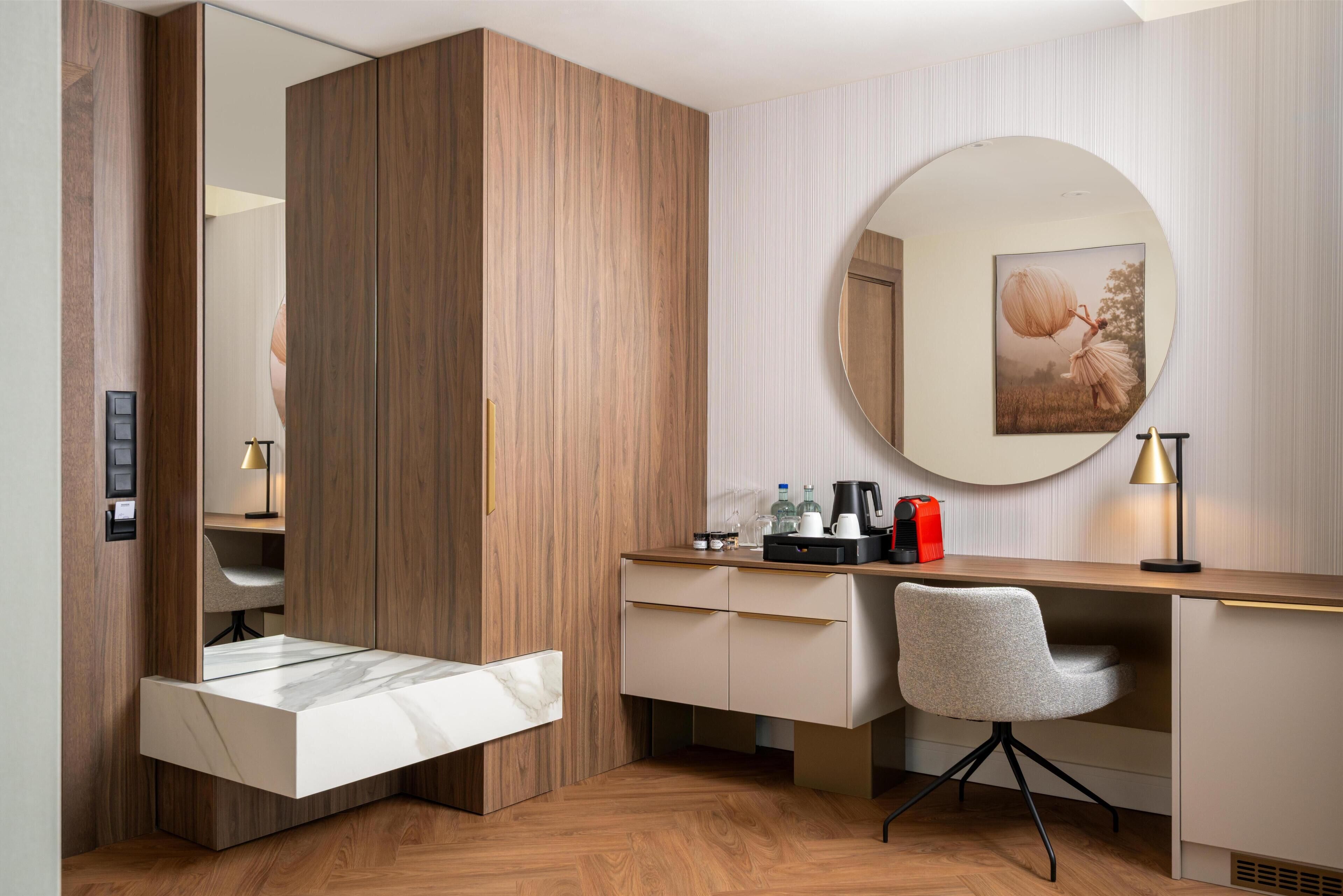Une chambre d’hôtel moderne, dotée d’une armoire en bois, d’un bureau avec une chaise, d’un miroir rond et d’un tableau encadré au mur.