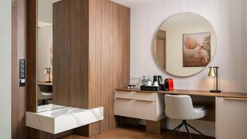 A modern hotel room with a wooden wardrobe, a desk with a chair, a round mirror, and a framed picture on the wall.