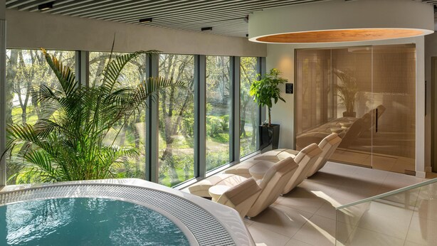 A modern indoor pool area with a view of a lush garden, a relaxation area with chairs, and a large window. - Radisson Blu Hotel, Cluj (Cluj-Napoca)