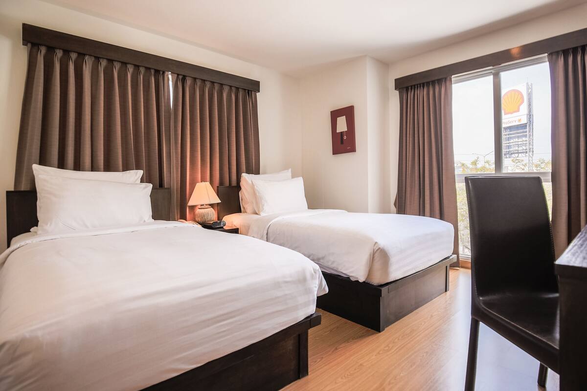 A hotel room with two beds, a desk, a chair, and a view of a cityscape.