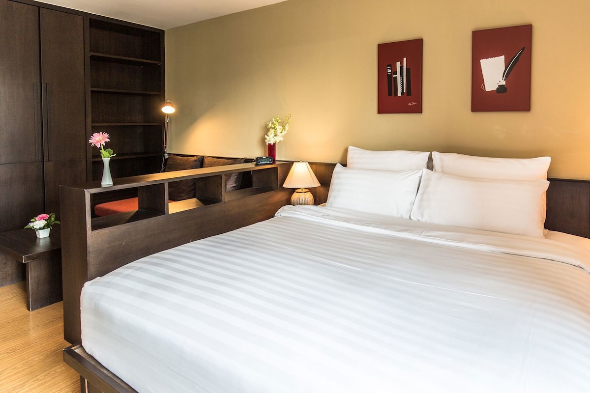 Quarto de hotel com cama grande, mesa de cabeceira com luminária, pequena mesa de cabeceira com um vaso de flores e prateleira na parede com um vaso.