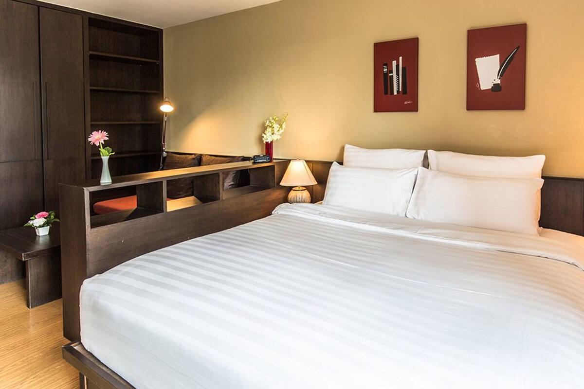 A hotel room with a large bed, a desk with a lamp, a small table with flowers, and two framed pictures on the wall.