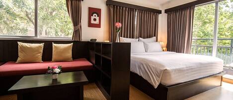 Una habitación de hotel moderna con una cama grande, un sofá rojo, una pequeña mesa con flores y ventanas amplias con cortinas.